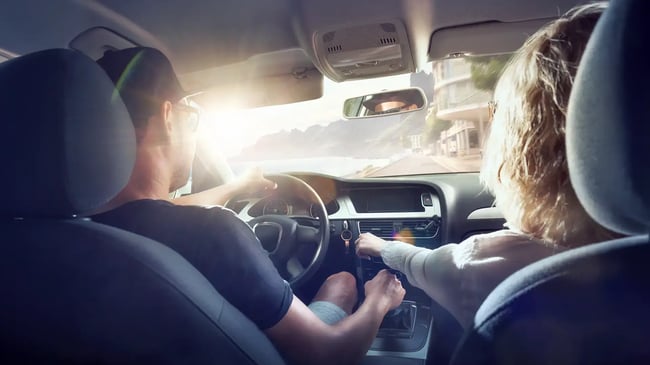 Zwei Personen sitzen auf den Vordersitzen eines Autos, das auf einer sonnigen Küstenstraße fährt. Durch die Windschutzscheibe sind die Straße, die Berge und das Wasser zu sehen.