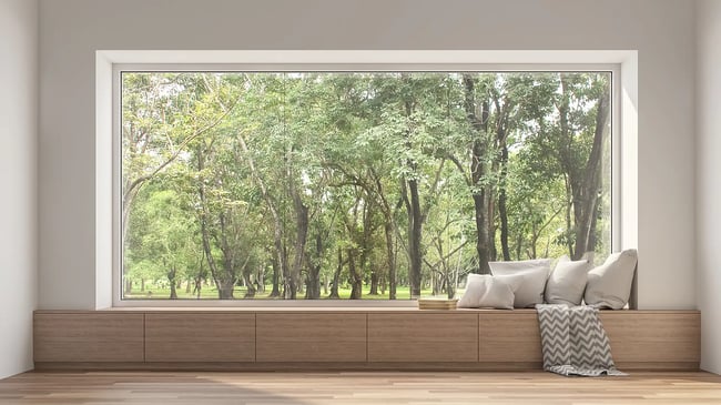 Ein Fenster mit einer eingebauten Holzbank und Kissen bietet einen Blick auf einen üppig grünen Wald. Sonnenlicht durchflutet den minimalistisch eingerichteten Raum mit seinen Holzböden und weißen Wänden.