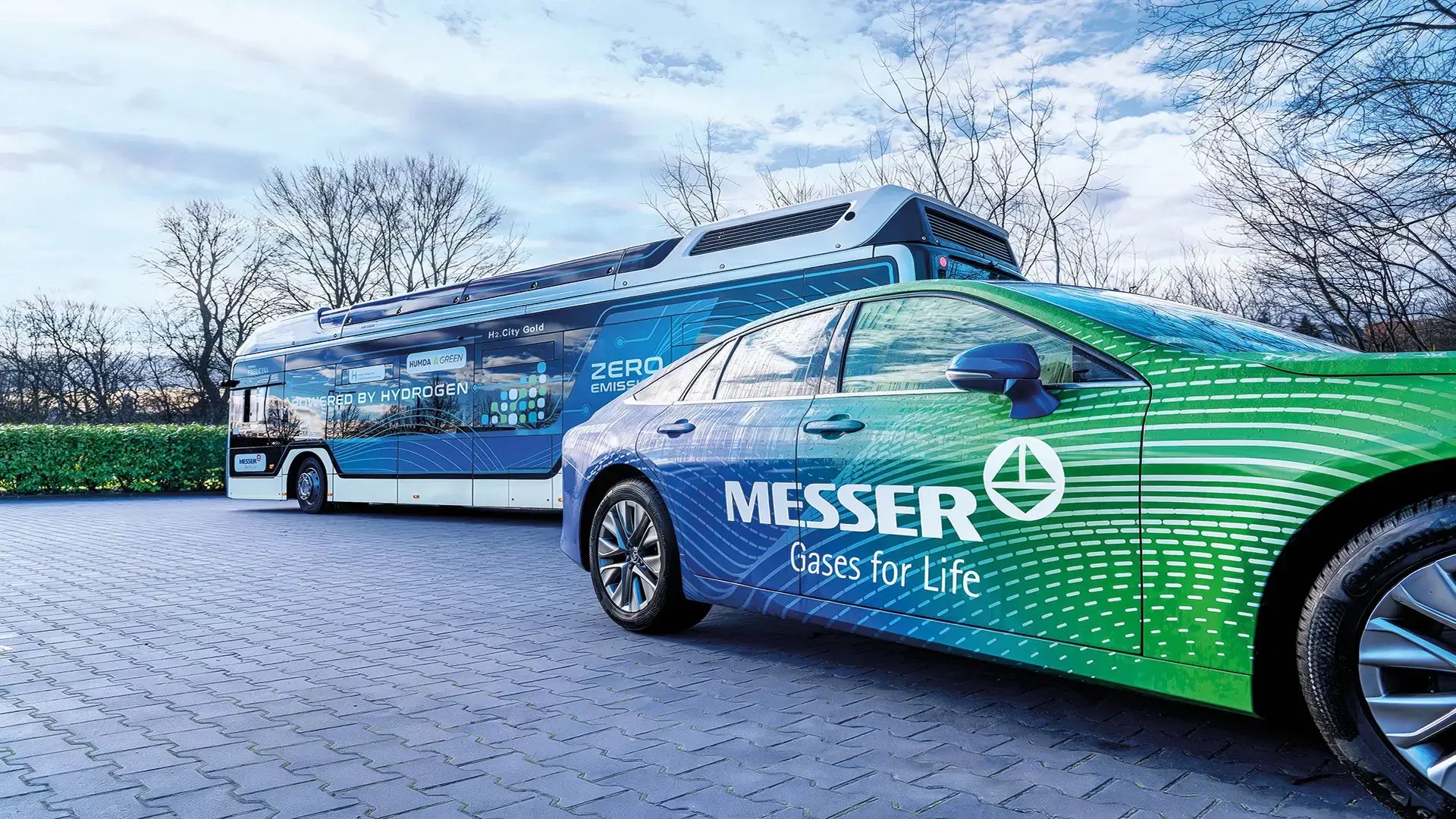 Ein grün-blaues Messer gebrandetes Auto, das mit Wasserstoff angetrieben wird, steht vor einem ebenfalls wasserstoffbetriebenen Bus unter einem teilweise bewölkten Himmel.