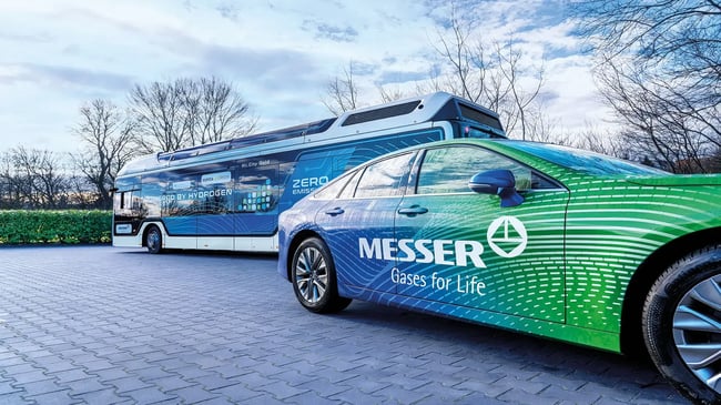 Ein grün-blaues Messer gebrandetes Auto, das mit Wasserstoff angetrieben wird, steht vor einem ebenfalls wasserstoffbetriebenen Bus unter einem teilweise bewölkten Himmel.