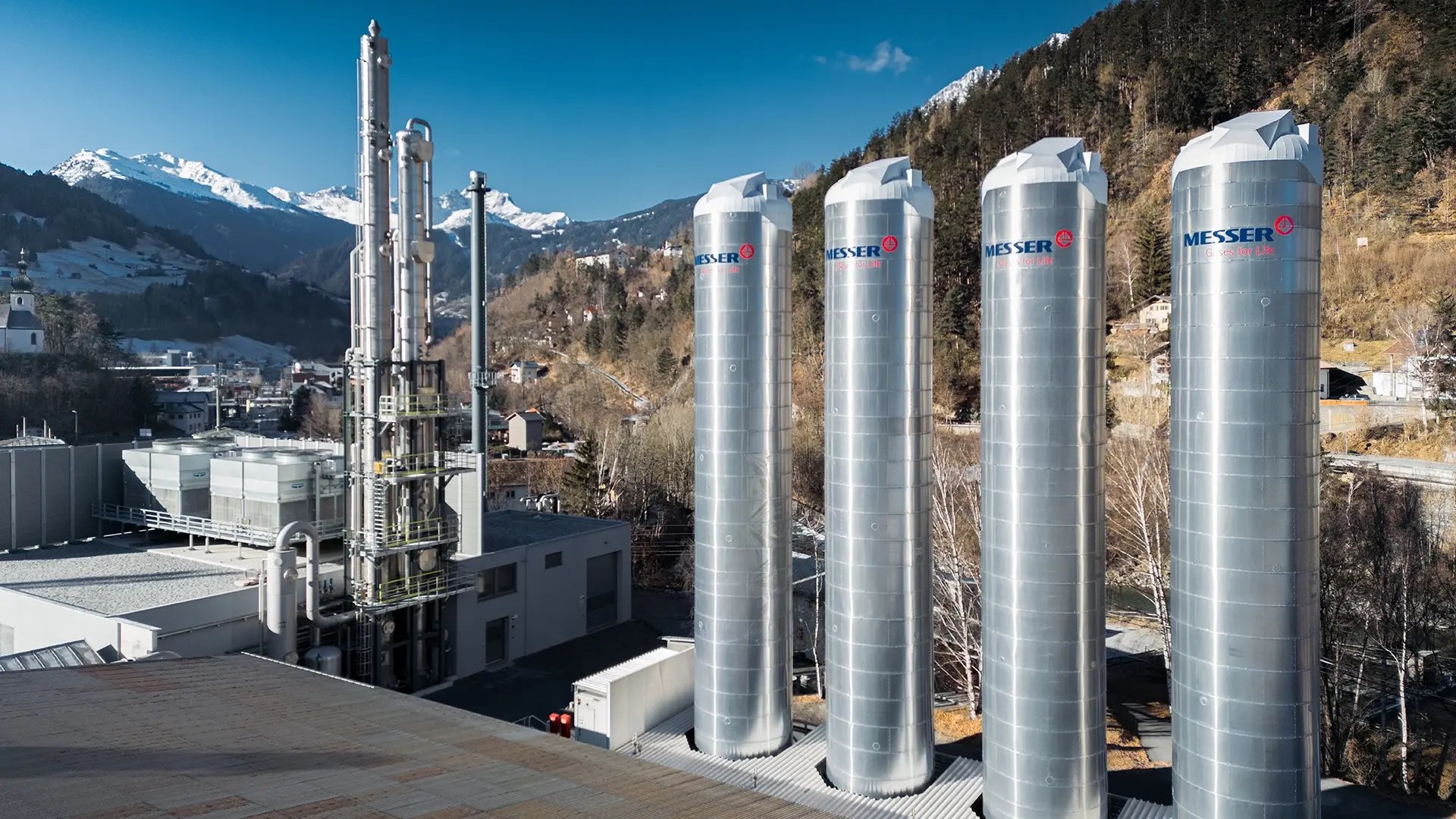 Four tall industrial gas storage tanks bearing the word “Messer” stand next to a carbon capture plant in a mountainous area under a bright blue sky.