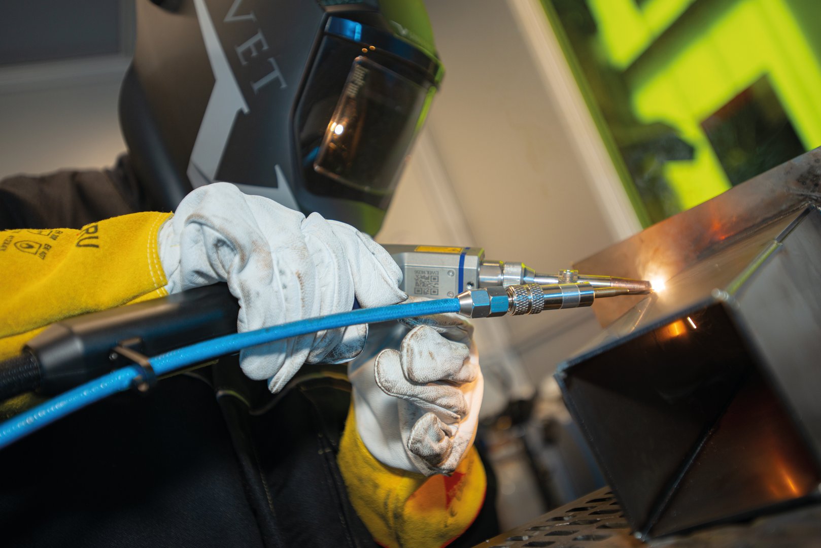 Ein Schweißer, der einen Schutzhelm und weiße Handschuhe mit gelben Bündchen trägt, verbindet in einer Werkstatt mit einem handgeführten Laserschweißgerät dunkle Metallteile miteinander.