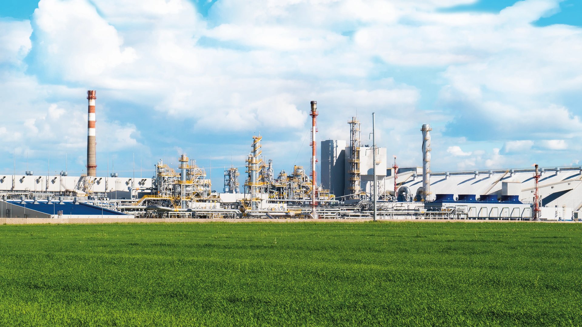 Industrieanlagen mit hohen Schornsteinen und der dazugehörigen Infrastruktur hinter einer grünen Wiese.