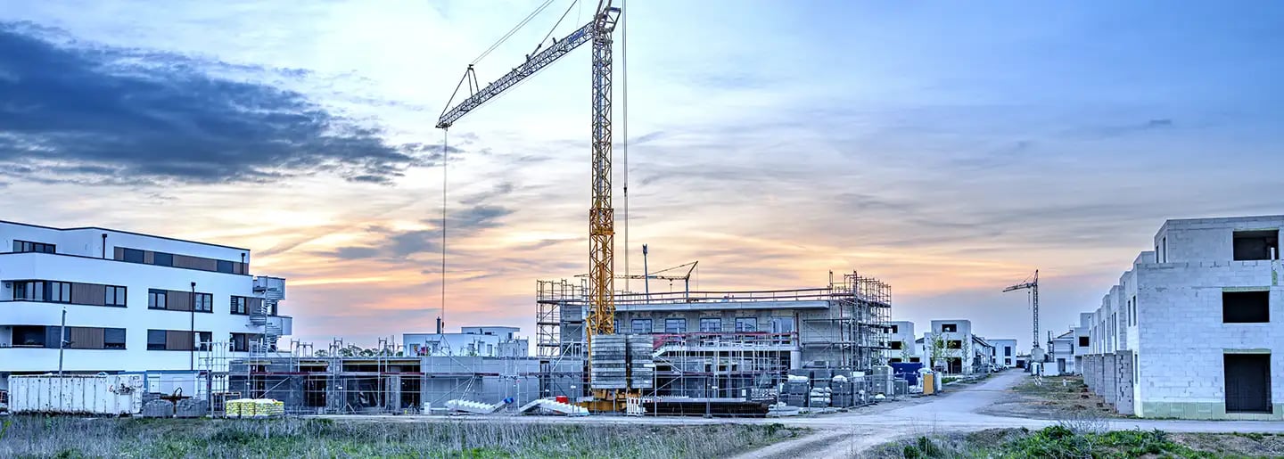 Baustelle bei Sonnenuntergang mit einem hohen gelben Kran, unfertigen Gebäuden, Gerüsten und modernen weißen Wohnblocks im Hintergrund unter einem teilweise bewölkten Himmel.