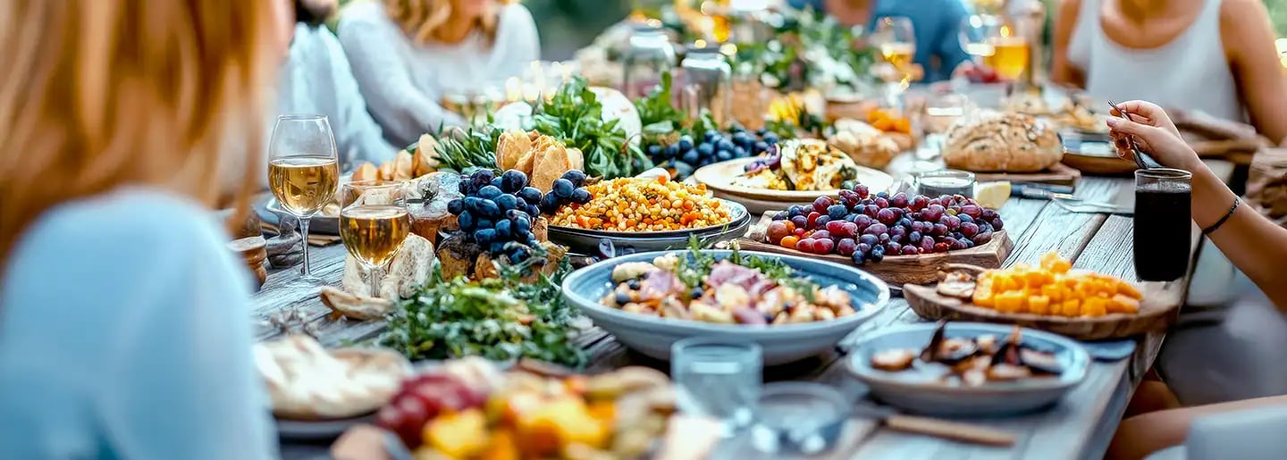 A group of people are sitting outdoors around a wooden table enjoying a colorful spread of food, including salads, bread, grapes, cheese, and drinks, creating a lively and festive atmosphere.