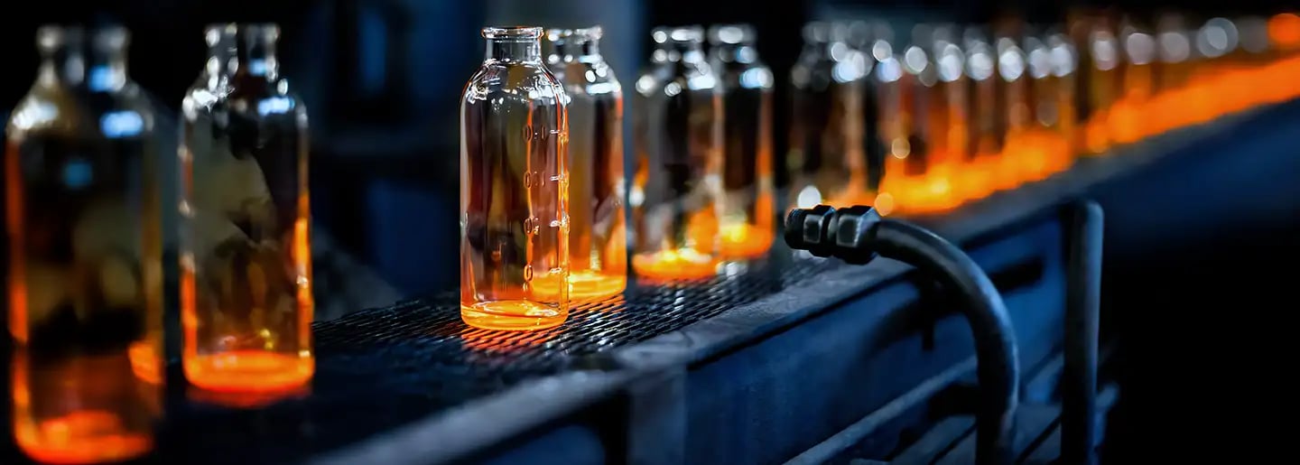 A row of glowing glass bottles on a conveyor belt in a factory. The bottles appear hot and orange, suggesting that they have just been molded or are being processed.