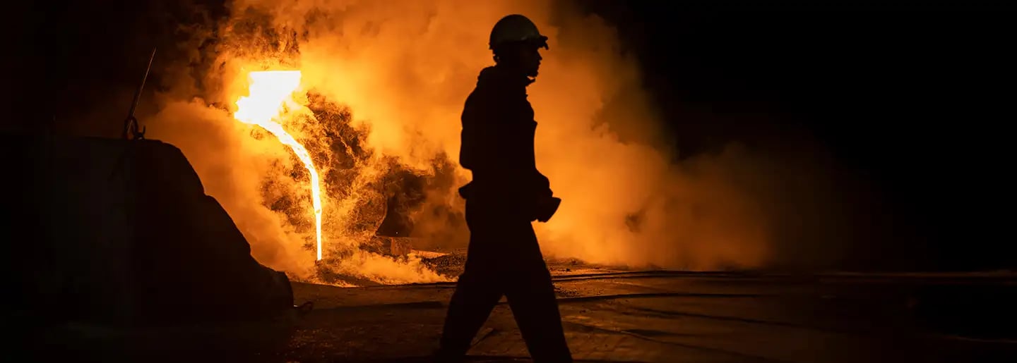 The silhouette of a worker stands in front of a glowing stream of molten metal flowing from a furnace. Thick steam and bright orange light illuminate the dark industrial scene.
