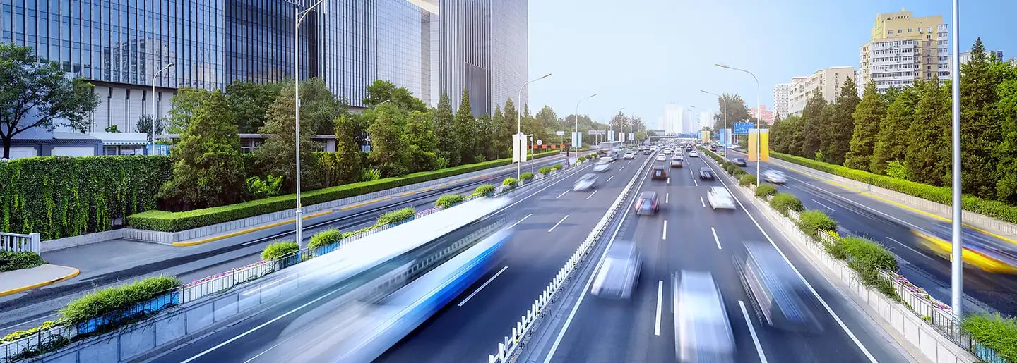 Eine vielbefahrene, mehrspurige Stadtautobahn mit verschwommenen Autos und Bussen in Bewegung, gesäumt von Bäumen und hohen, modernen Bürogebäuden unter einem klaren, blauen Himmel.