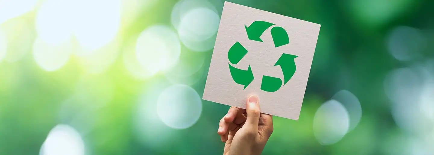 A hand holds up a white card with a green recycling symbol in front of a blurred natural background with shades of green and yellow.